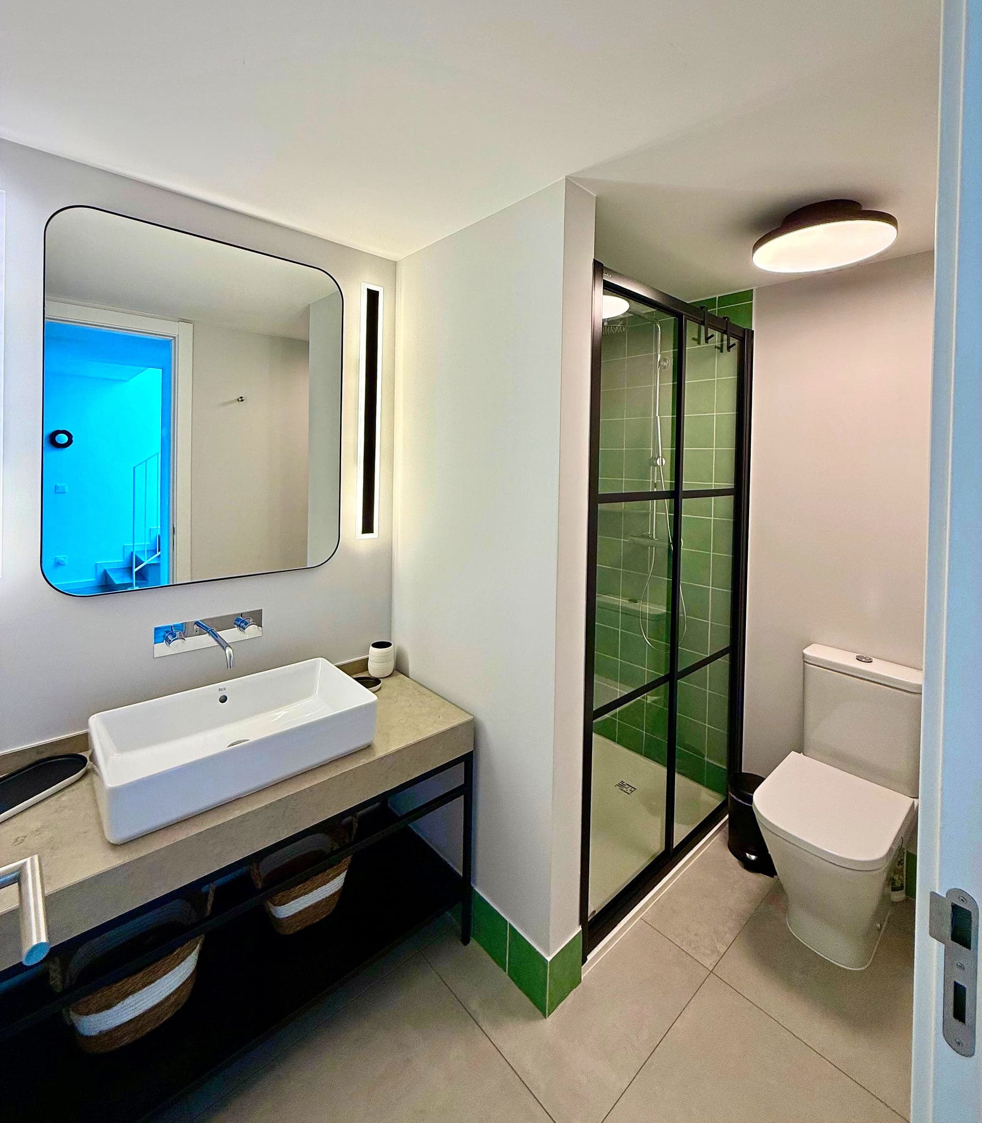 Casa de banho com lavatório branco sobre bancada de pedra, espelho com luz, duche de chão com azulejo verde e sanita