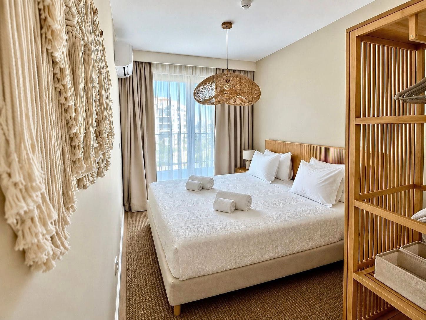 Cama de casal com roupa branca e toalhas enroladas, um macramé na parede, candeeiro de ratã e roupeiro de ripas