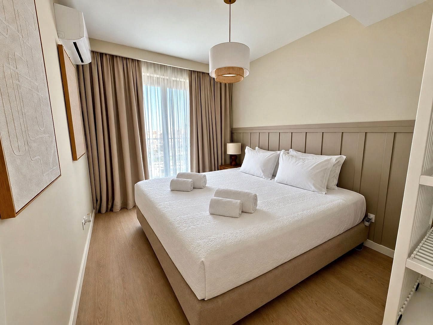 Cama de casal feita, com toalhas, parede em lambrim, candeeiro de tecido e uma janela alta com cortinas